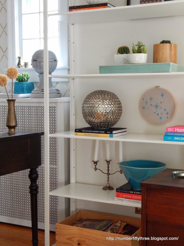 White shelving unit with decorative objects including a silver globe, books, candlestick, blue bowl, an easy DIY beginner embroidery project in a frame, and a wooden crate; partial view of a desk in the foreground.