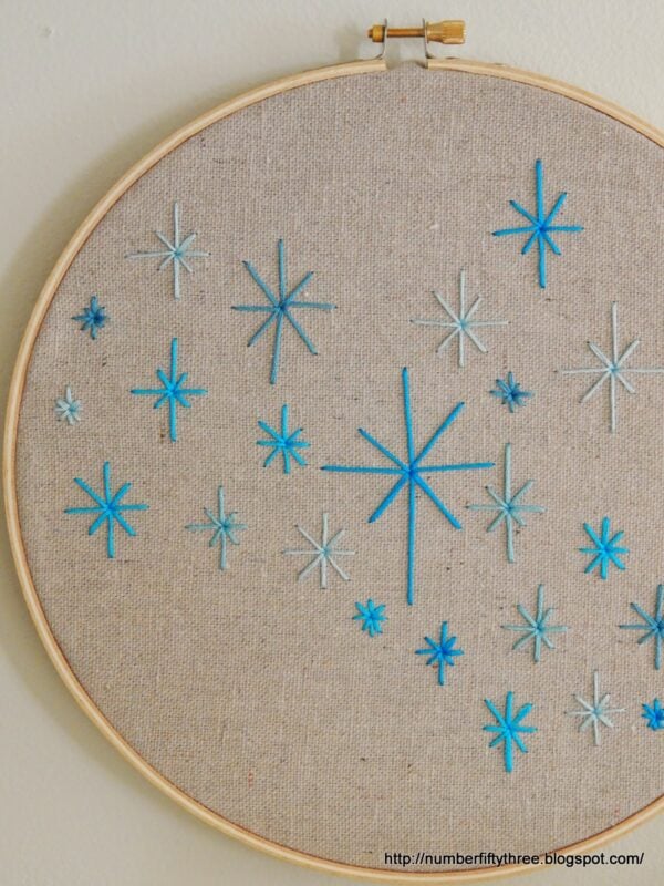 An easy DIY beginner embroidery project featuring an embroidery hoop with various sizes of blue and teal starburst designs on beige fabric.