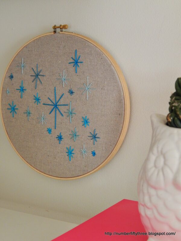 A wooden embroidery hoop displayed on a wall features various blue starburst patterns stitched on beige fabric—an easy DIY beginner embroidery project. A white ceramic vase is partially visible in the foreground.