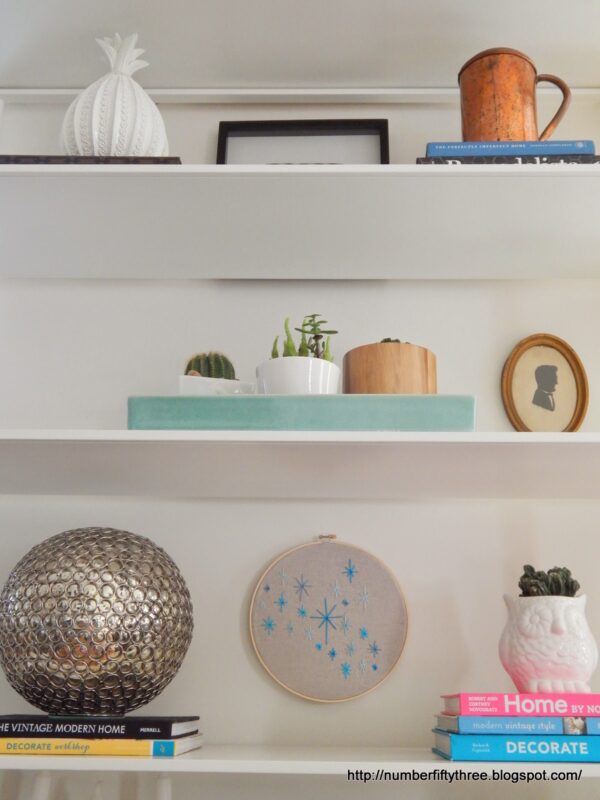 White shelves display a mix of decor items, including books, a silver sphere, potted plants, a framed silhouette, copper mug, pineapple-shaped jar, and an easy DIY beginner embroidery project in a blue-embroidered hoop.
