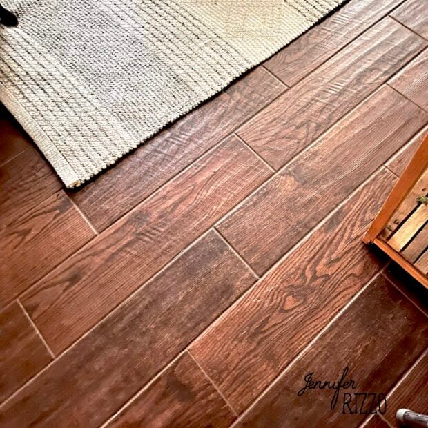 A close-up view of a floor with brown wood look ceramic tile, a beige woven rug, and a wooden furniture piece in the corner.