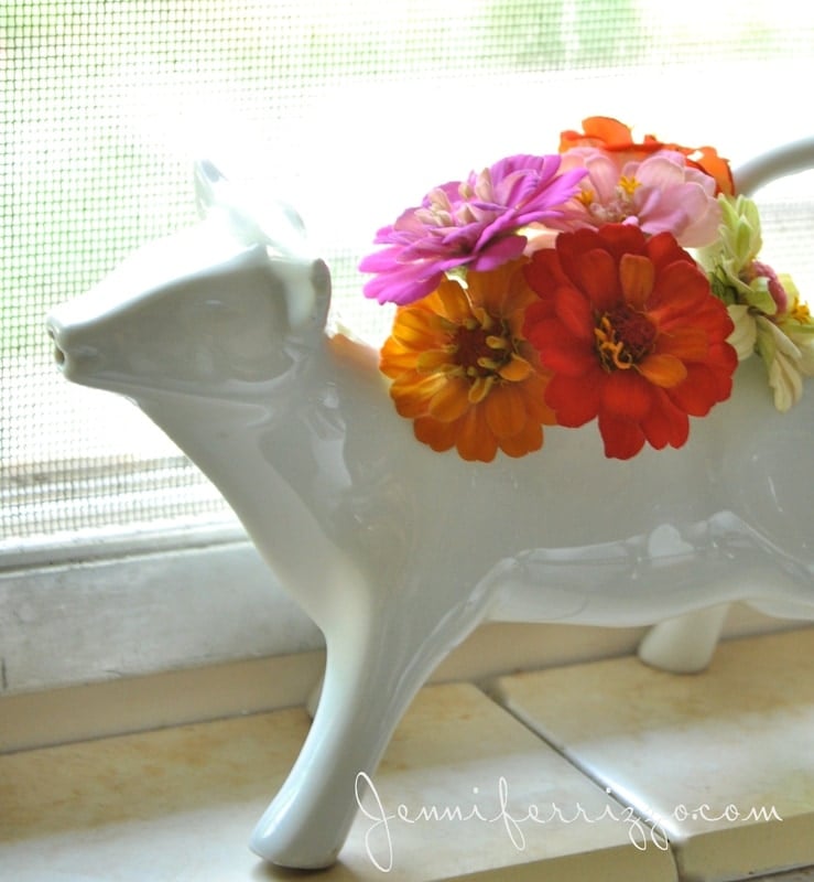 A white ceramic cow-shaped vase, looking like one of those charming repurposed vintage animal gravy boats, holds colorful flowers on a windowsill.