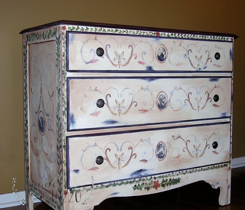 Wooden dresser with three drawers, featuring ornate, hand-painted floral and vine designs in pastel colors. This stunning dresser makeover sits against a plain wall on a hardwood floor.