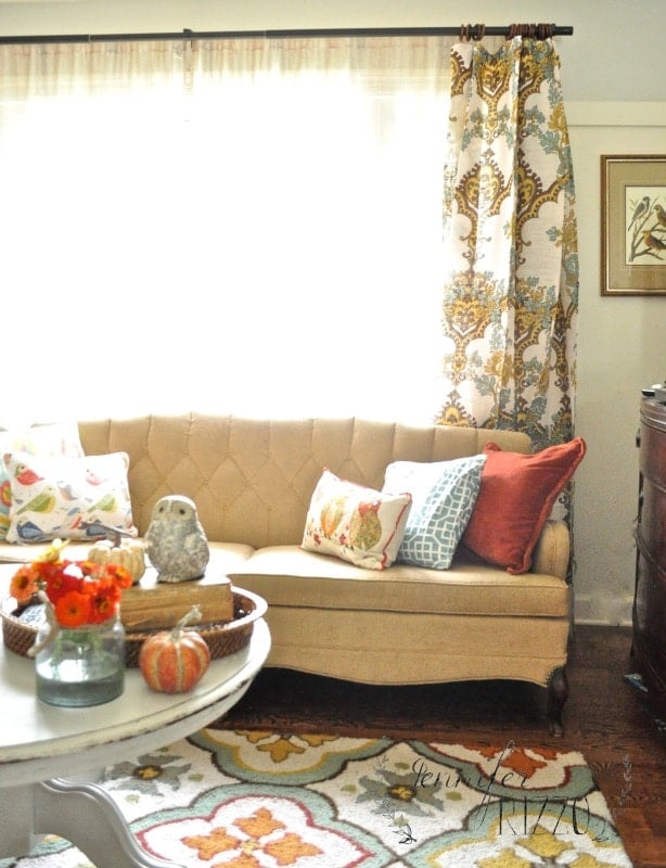 A beige tufted sofa with assorted colorful throw pillows sits in front of a window with patterned curtains in a bright, cozy living room, ready for a Fall home tour. A round table with autumn-inspired decor is in the foreground.