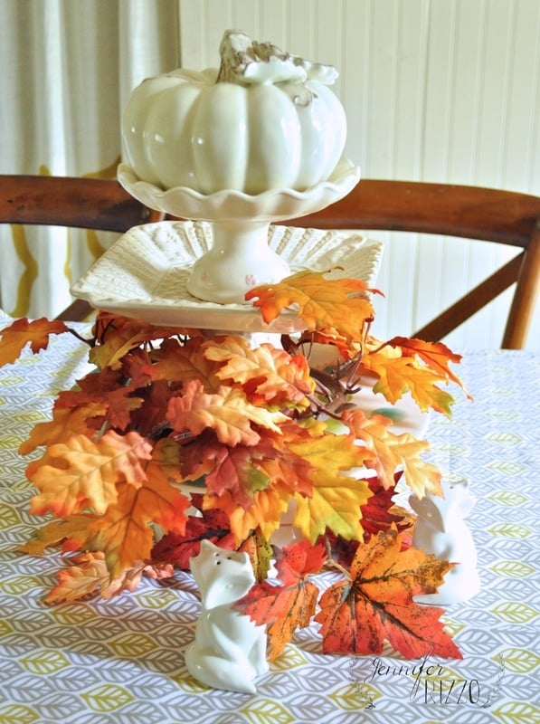 On a fall home tour, a white ceramic pumpkin sits elegantly on a pedestal, surrounded by a cascade of autumn leaves and charming white squirrel figurines atop a patterned tablecloth.
