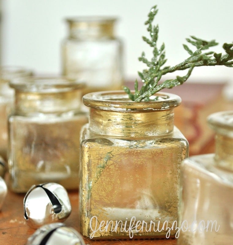Several small, square DIY gold antiqued glass jars with gold trim are arranged on a surface, one holding a green twig. Silver jingle bells are scattered nearby.