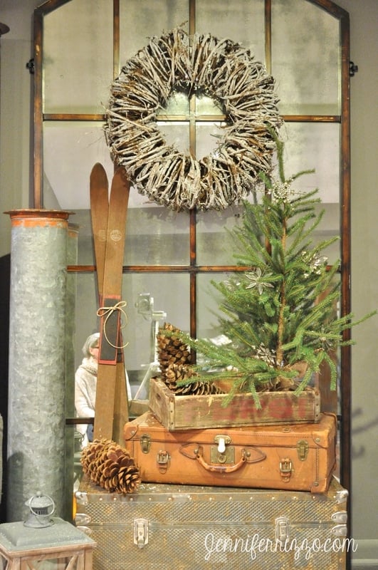A small pine tree, pinecones, vintage suitcases, and skis are arranged in front of a window with a rustic wreath above—perfect inspiration for cozy Basement Christmas Decorating Ideas.