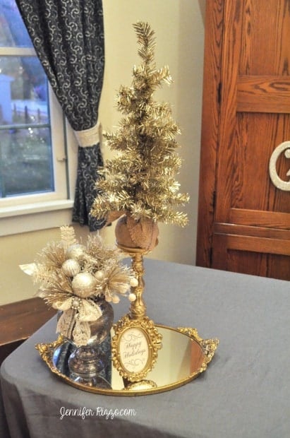 A creatively Christmas display with a small gold tree, a vase of festive ornaments, and a framed "Happy Holidays" message on a golden tray, positioned on a table near a window with a blue curtain.