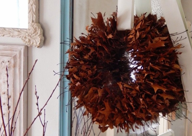 An oak leaf wreath made of dried brown leaves hangs on a white ribbon against a mirror, with a light-colored frame and branches in the background.