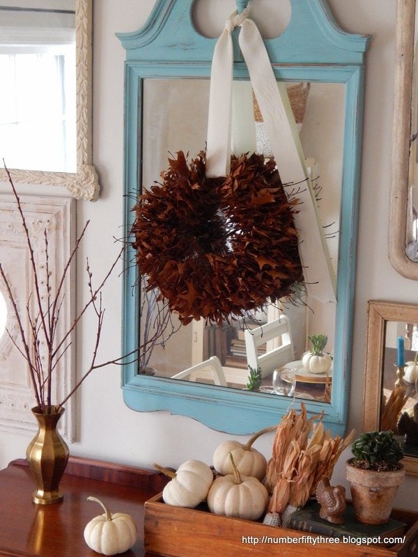A decorative oak leaf wreath hangs on a blue mirror, with white mini pumpkins and a gold vase with twigs placed on a wooden dresser below.