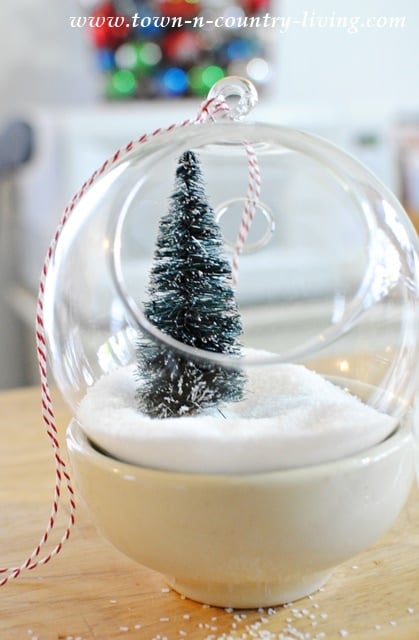 A DIY hanging snow globe ornament featuring a miniature snow-covered tree and artificial snow sits in a white bowl on a wooden surface.