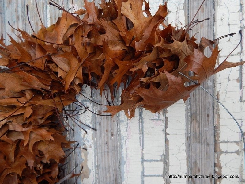 A close-up view of an oak leaf wreath made of dried brown leaves against a weathered wooden background with peeling paint. A blog URL is visible in the bottom right corner.