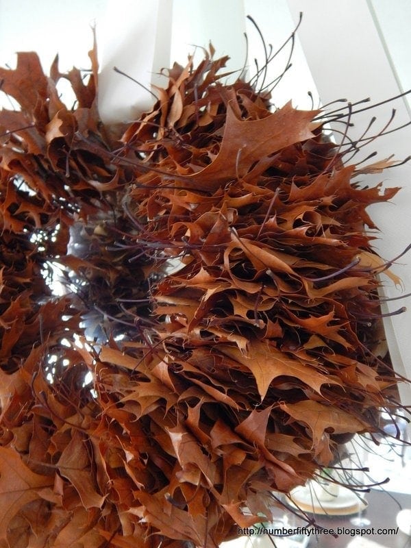 An oak leaf wreath made from brown dried leaves is displayed hanging on a white wall.