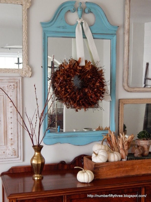 A blue-framed mirror with an oak leaf wreath hangs above a wooden sideboard. The sideboard has a brass vase with twig branches and several white pumpkins.