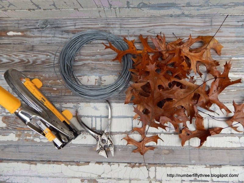A bundle of wire, dried oak leaves, pruning shears, and a stapler are arranged on a wooden surface, ready for crafting an elegant oak leaf wreath.
