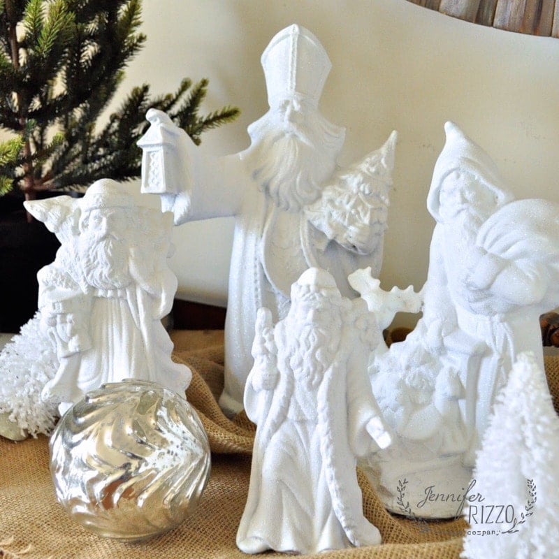 Upcycled ceramic thrift store Santas of Santa Claus and St. Nicholas are arranged on a burlap surface, with a silver ornament and a small pine tree in the background.