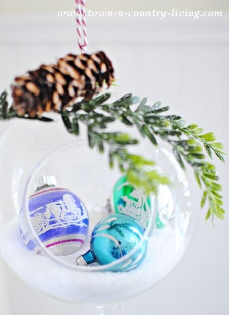 A glass filled with white artificial snow, three colorful Christmas ornaments, and decorated with a pinecone and green foliage hanging above—perfect inspiration for DIY hanging snow globe ornaments.