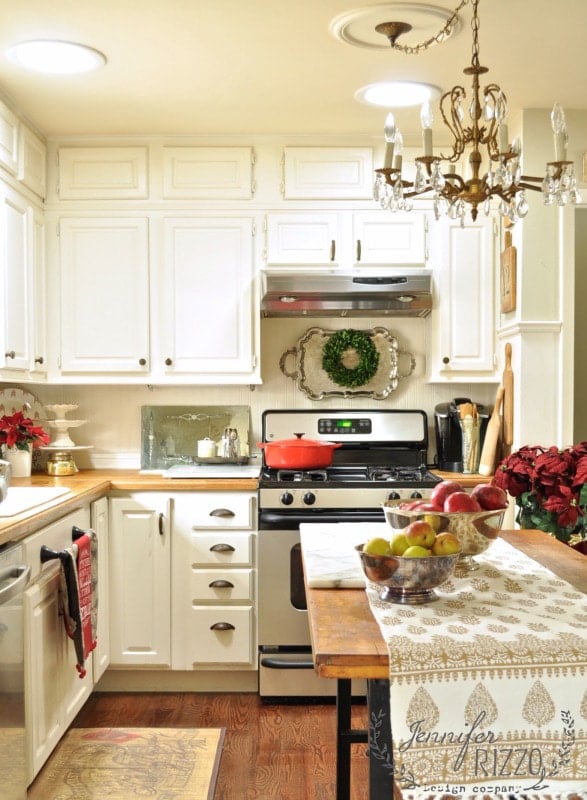 A small kitchen with white cabinets, a stove with a red pot, a wooden table with a fruit bowl, festive holiday decorations, and a chandelier overhead—perfect for enjoying a holiday house walk.