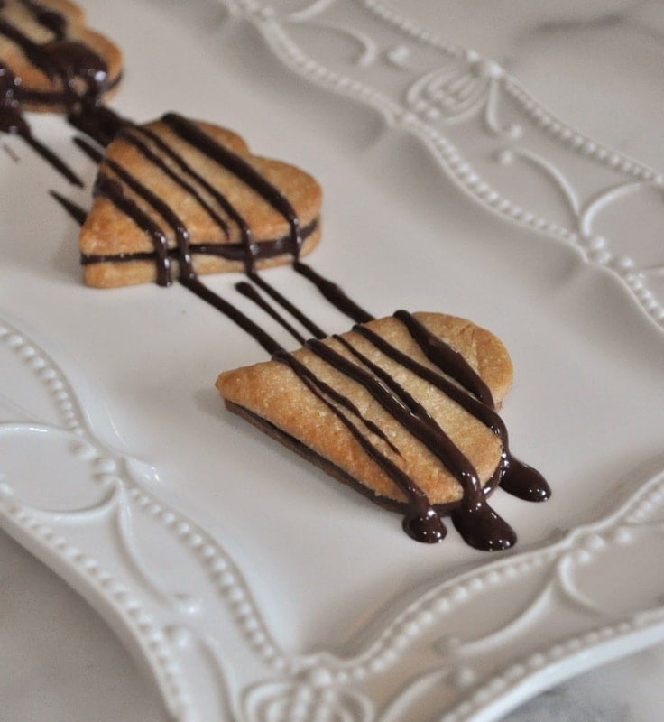 Three heart-shaped pie crust cookies drizzled with chocolate sauce are arranged in a line on a decorative white serving platter.