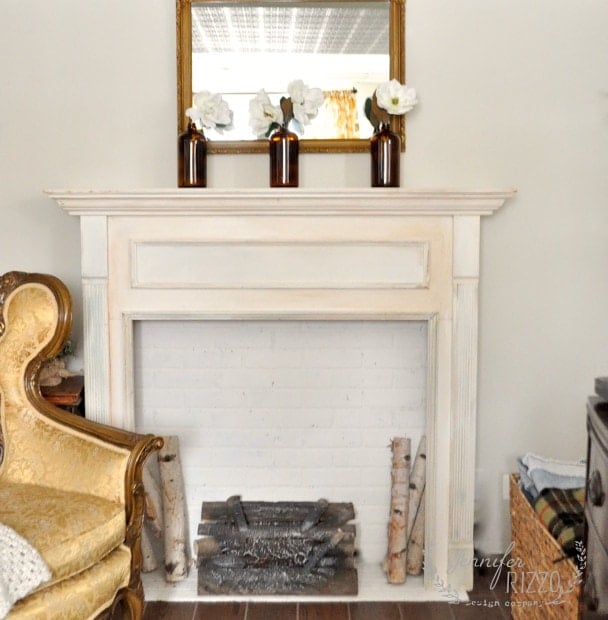 A decorative faux brick fireplace with a mantel displaying three vases of white flowers. An ornate armchair is positioned to the left, while logs and a grate sit elegantly in the white fireplace opening.