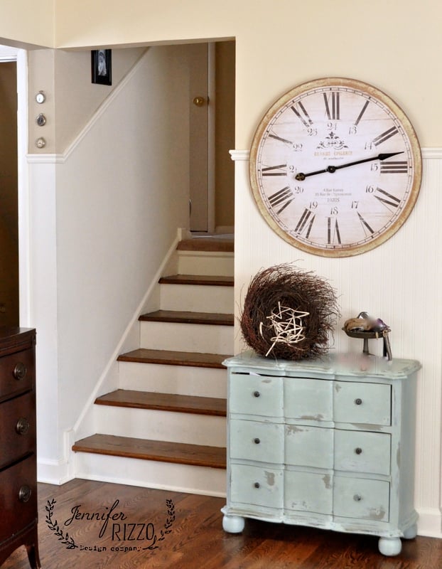 A vintage-style clock hangs above a distressed light blue dresser, offering charming furniture with storage for decorative items, next to a wooden staircase in a cozy home interior.