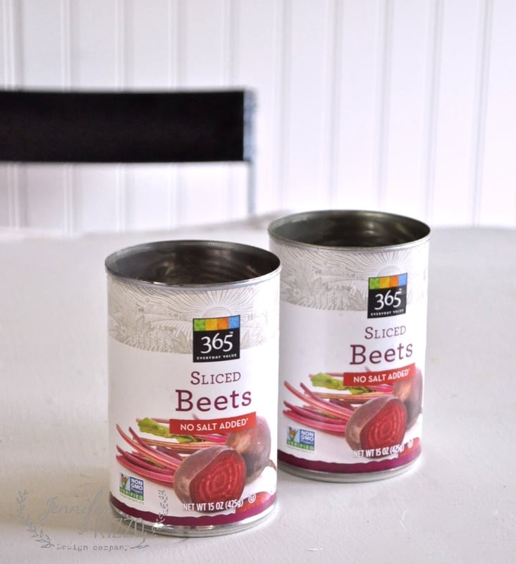 Two cans of 365 brand sliced beets labeled "No Salt Added," rest on a white surface. Their shape and size resemble cute containers for battery operated candles, with a sleek black chair subtly positioned in the background, adding contrast to the scene.