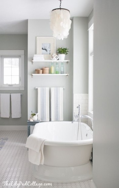 Bathroom with a freestanding tub, light gray walls opening up your space with paint color, a window, a chandelier, and shelves holding plants and toiletries.