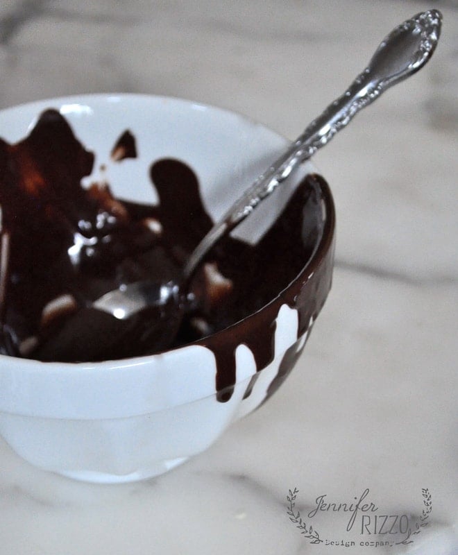 A white bowl with remnants of melted chocolate inside and a spoon placed in it, sitting on a marble surface. The credit "Jennifer Rizzo" is visible at the bottom right. Nearby, there's a hint of pie crust cookies adding to the delectable scene.