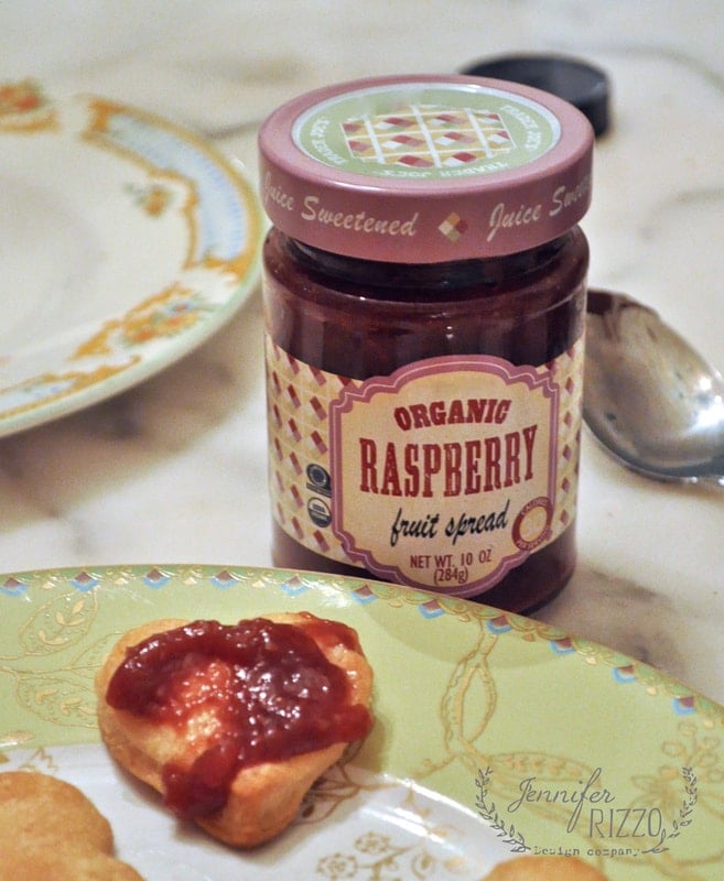 A jar of organic raspberry fruit spread is on a table next to a spoon, a piece of bread topped with the spread, and some pie crust cookies.
