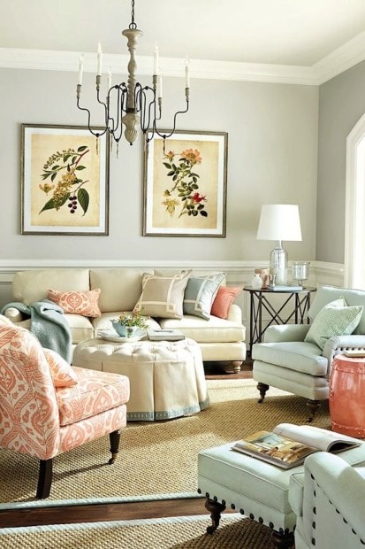 A cozy living room featuring neutral-toned sofas and floral prints on the wall, thoughtfully arranged with an eye for furniture placement around windows. A round ottoman sits under a chandelier, while decorative cushions brighten the seating area.