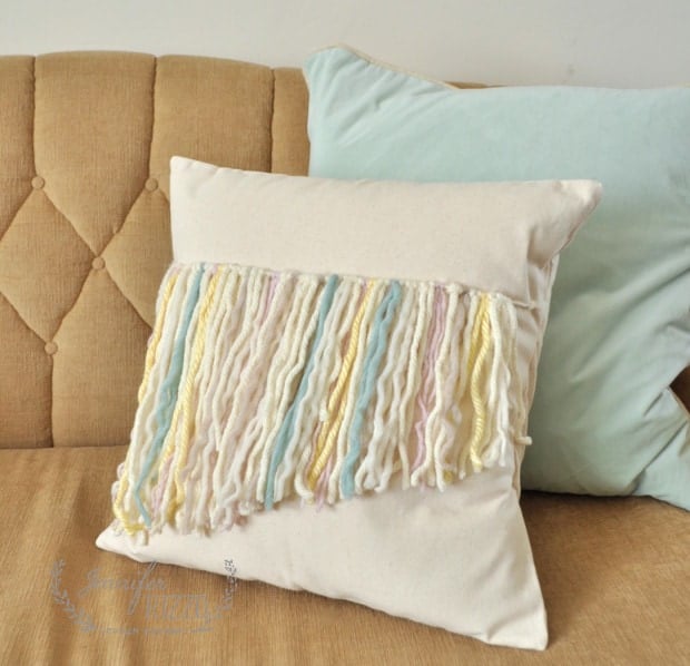 A beige pillow with pastel-colored yarn fringe sits on a tan cushioned couch, accompanied by a light blue pillow.