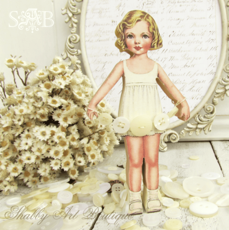 Vintage button craft comes to life as a paper doll in a white dress stands among white flowers and buttons, framed by a decorative border and handwritten script in the background.