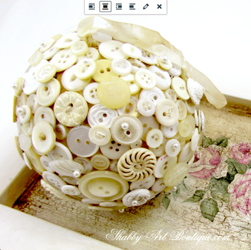 A decorative sphere, crafted using the vintage button craft technique and covered with assorted white and cream buttons, rests on a vintage-style tray adorned with floral artwork.