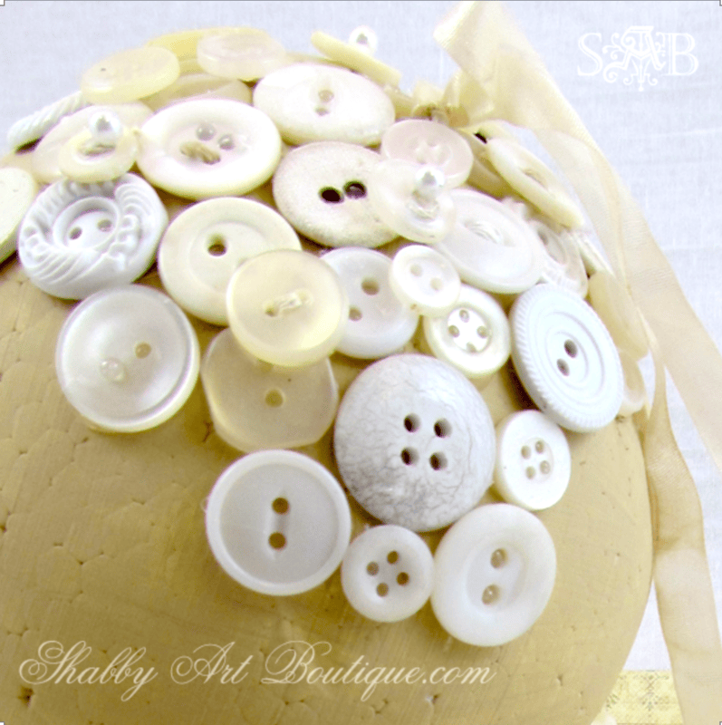 A collection of white and cream buttons is arranged on a textured, light yellow surface, evoking the charm of vintage button craft, with a sheer ribbon visible in the background.