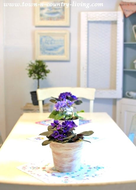 A white table with potted purple flowers placed on vintage handkerchiefs, centered in a bright room with framed art and a small plant in the background.