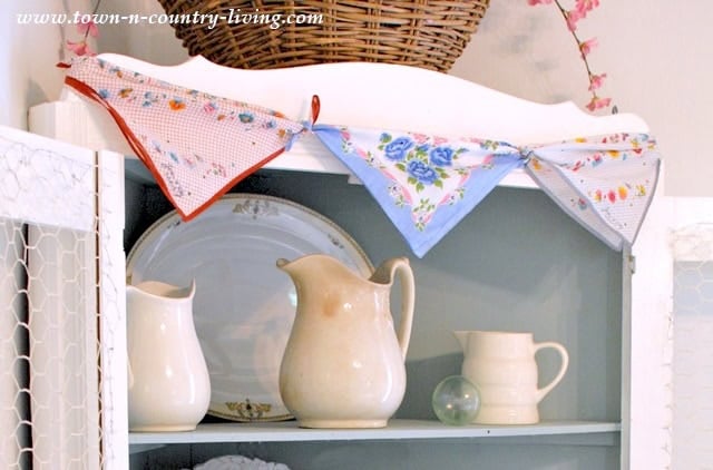 A white cabinet displays cream-colored pitchers and plates, with a string of vintage handkerchiefs hanging across the top.