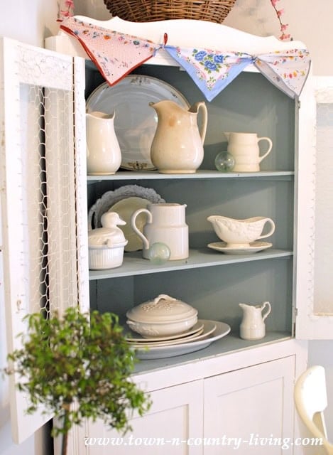 A white corner cabinet with chicken wire doors displays cream-colored vintage pitchers, plates, and serving dishes on blue shelves; vintage handkerchiefs, a basket, and linens are arranged on top.