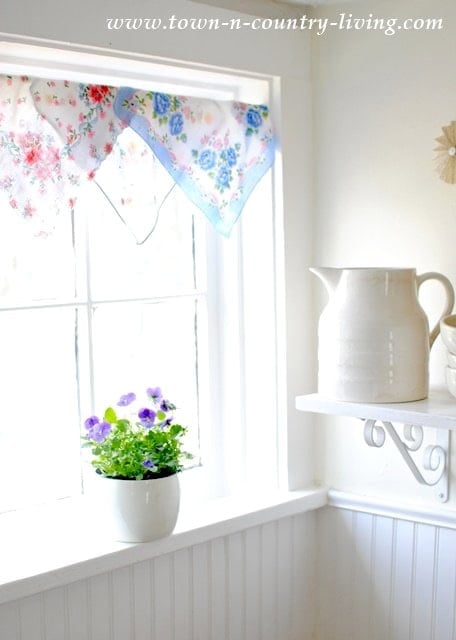 A small potted plant with purple flowers sits on a windowsill beside a white pitcher and folded vintage handkerchiefs on a wall shelf in a bright, white room.