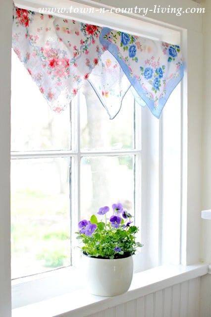 A white windowsill with a potted purple pansy plant sits below a window, which is decorated with vintage handkerchiefs featuring floral patterns as a charming valance.