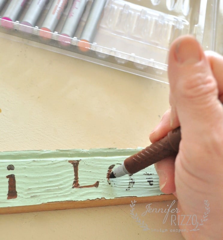 A hand holding a brown pen writes "LOVE" on what appears to be a vintage wood sign, with its textured surface adding charm to the scene. In the background, a plastic container filled with colorful markers sits quietly, waiting for their moment of creativity.