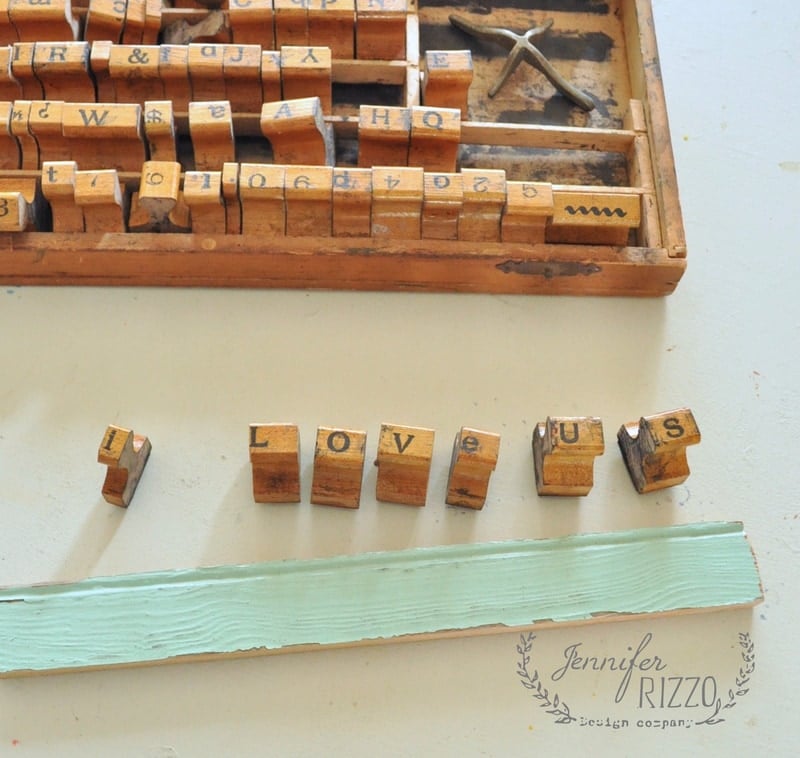 Wooden letter stamps spell "love us" beside an aqua strip of wood, evoking the charm of a vintage wood sign, with additional letter blocks in a tray above.
