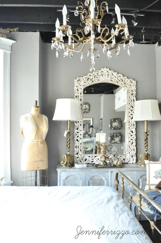 Elegant bedroom decor featuring a vintage mannequin, ornate mirror, chandelier, and two lamps on a dresser. A portion of a bed is visible in the foreground.