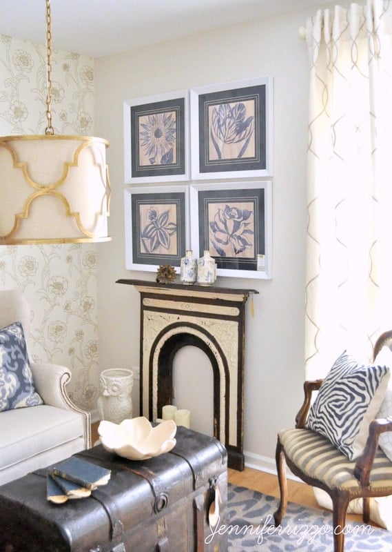 A cozy living room harmoniously blends modern traditional elements, featuring a white sofa, patterned armchair, trunk-style coffee table, wall art, ornate fireplace, and decorative lamp.