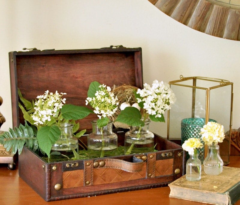 An open vintage suitcase reveals an array of glass vases filled with white flowers and ferns. Nearby, smaller glass bottles hold delicate blooms alongside a gold candle holder with a tealight.