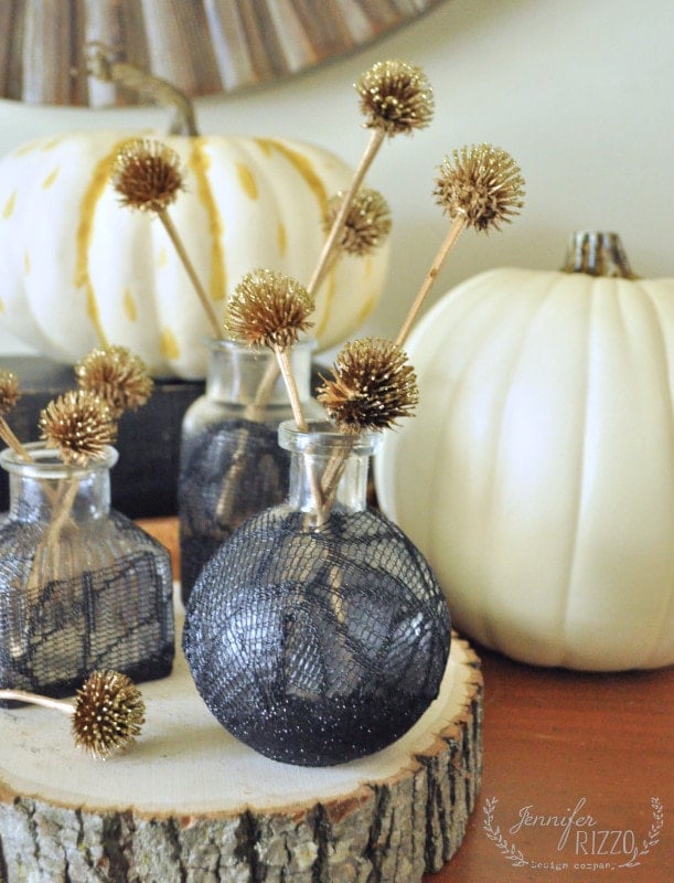 A DIY touch adds charm as decorative white pumpkins and black vases with metallic gold pom-pom sticks are elegantly arranged on a wooden slice. Decoupage black lace Halloween bottles further accentuate the festive display, creating a chic and spooky ambiance.