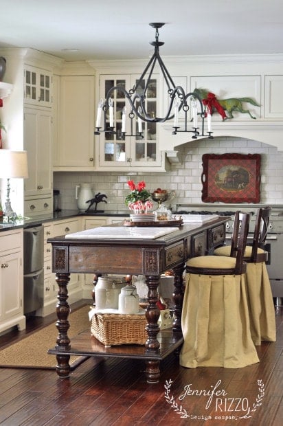A traditional kitchen in a converted horse barn to amazing home, with white cabinets, a dark wood island, skirted barstools, a chandelier, and festive holiday decorations.