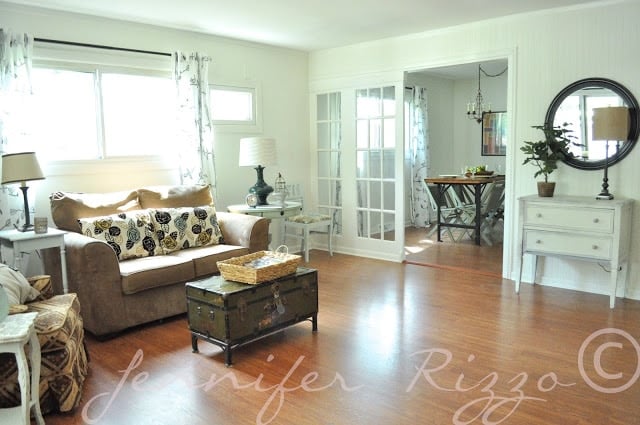 A bright living room in a charming ranch house, featuring a brown sofa, patterned pillows, and a wooden chest as a coffee table. Glass doors lead to the dining area with a round table and chairs, offering inspiration for those considering remodeling ideas.