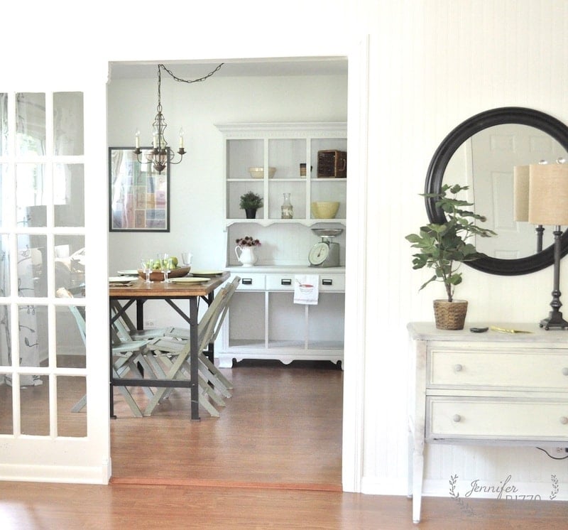 A dining room with a wooden table and chairs, white hutch, and decorative items complements this ranch house remodeling idea. Adjacent is a hallway featuring a round mirror and a small dresser adorned with a lamp and plant.