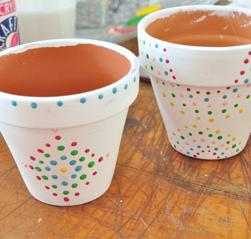 Two paint dot flower pots—terracotta and painted white with colorful spots—sit cheerfully on a wooden surface.