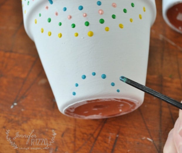 A hand uses a thin brush to paint blue dots on the bottom of a white flower pot, adding to its cheerful paint dot flower pots design with colorful polka dots.